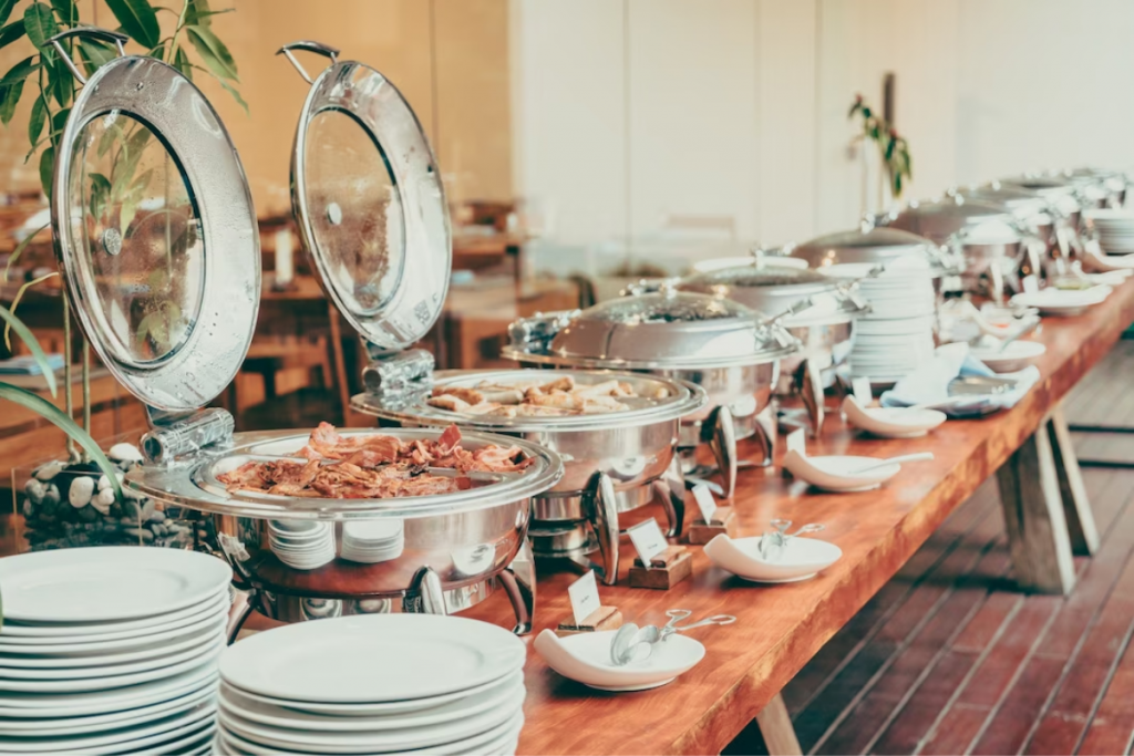 Buffet disposto em mesa de madeira com estações de metal que contém comida, em um serviço de catering. 